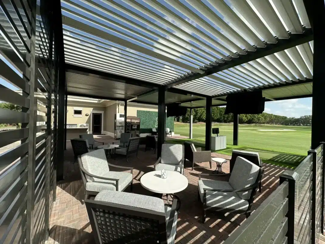 Outdoor lounge area under a slatted pergola with gray seating and white tables, overlooking a grassy golf course.