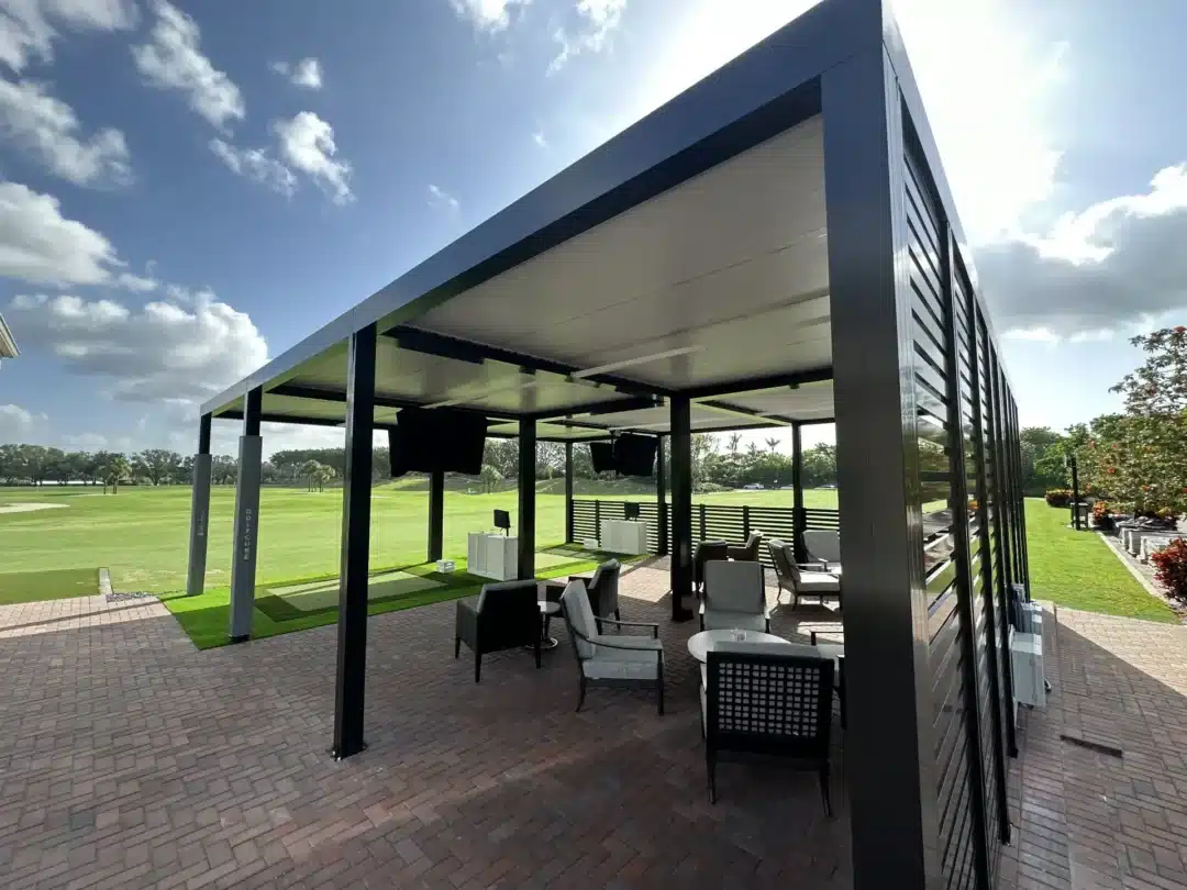 Outdoor covered seating area with black metal frame, wicker chairs and tables on a brick patio, overlooking a green golf course.
