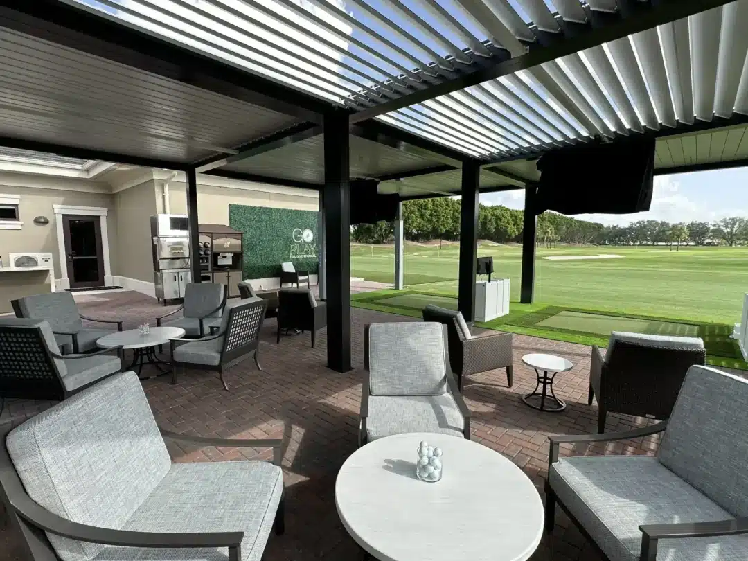 Outdoor covered patio with gray cushioned lounge chairs and white round tables, overlooking a green golf course.