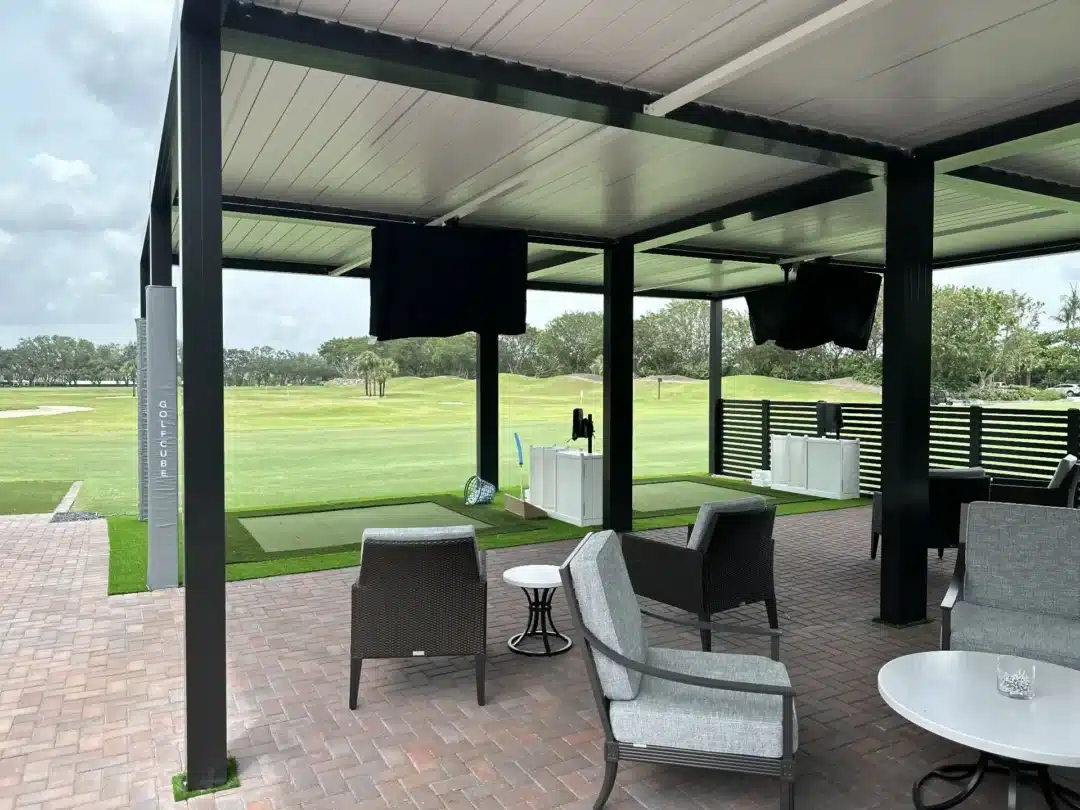 Covered outdoor seating area with gray wicker chairs and white tables on brick patio, facing a golf course. t