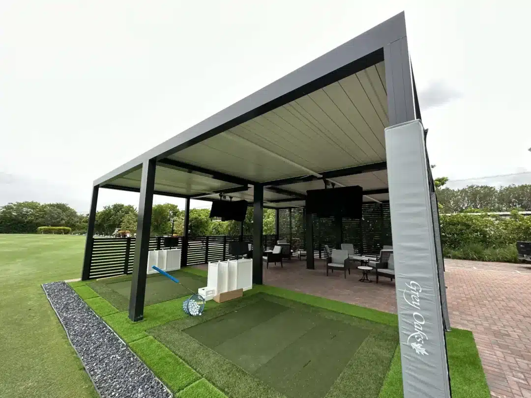 Open-air modern pavilion with a black metal frame, seating area, and artificial turf mats for golf practice under a large roof.