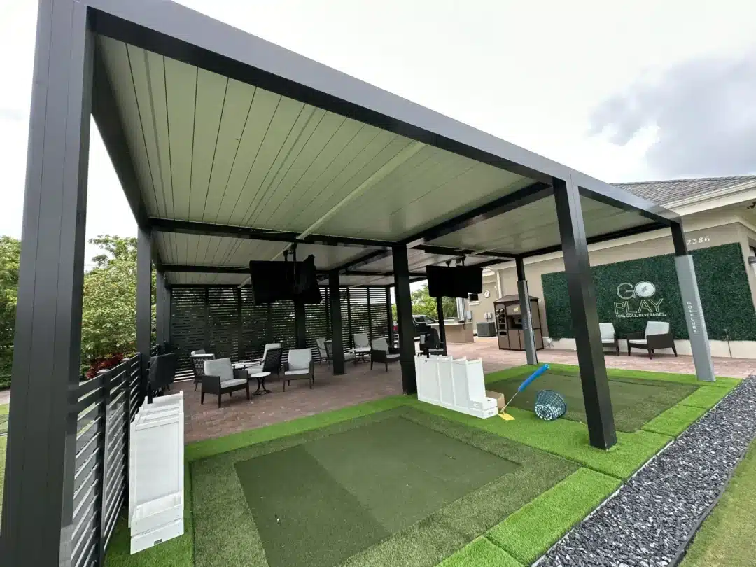 Outdoor lounge area under a dark metal pergola with chairs, tables, and hanging TVs, next to a Go Play sign on a green wall.