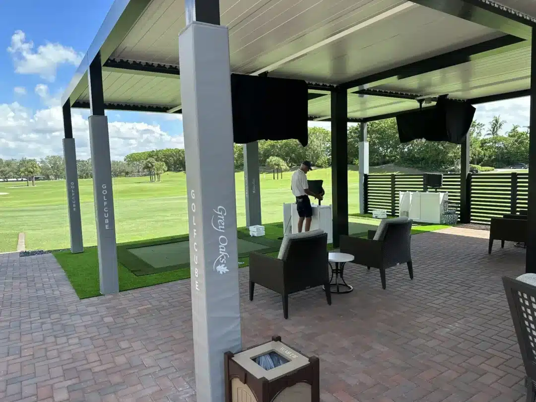 Covered outdoor seating area at a golf club with banners on tall posts and wicker chairs overlooking a green lawn