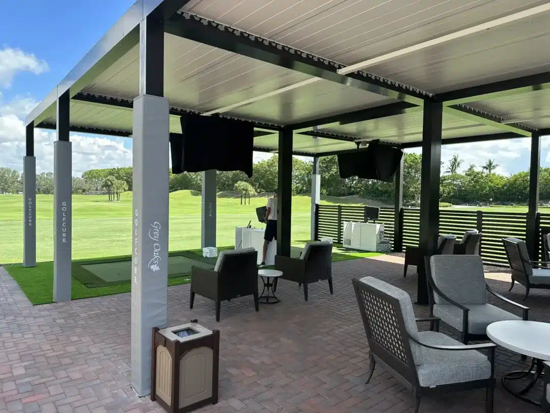 Outdoor golf club terrace with shaded seating under a metal roof, black fabric dividers, and green lawn beyond the patio.