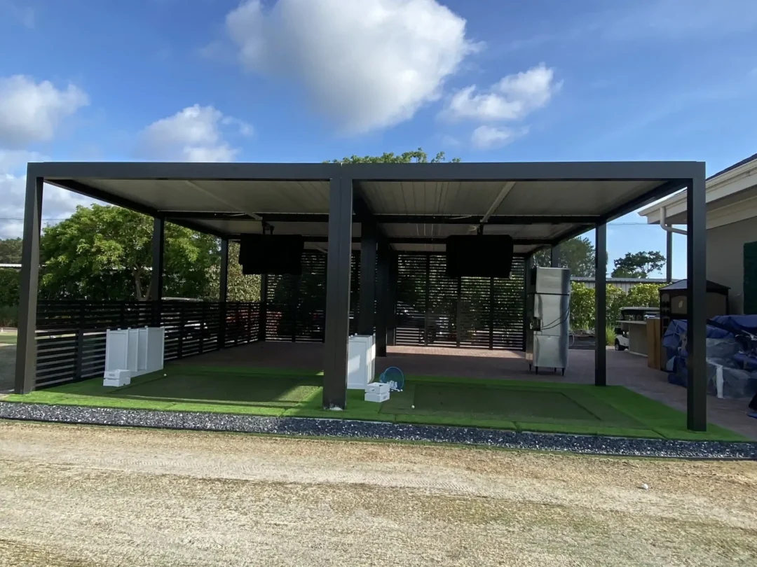 Modern dark gray metal carport/pavilion with flat roof and open sides over a paved area, artificial turf, and scattered construction pieces nearby under a blue sky with clouds.