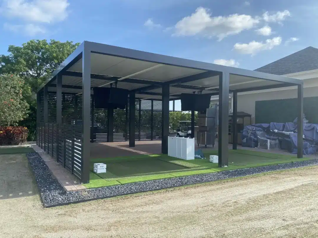 Modern black metal pergola with slatted walls, creating an outdoor covered area in a backyard, with covered furniture and equipment nearby.