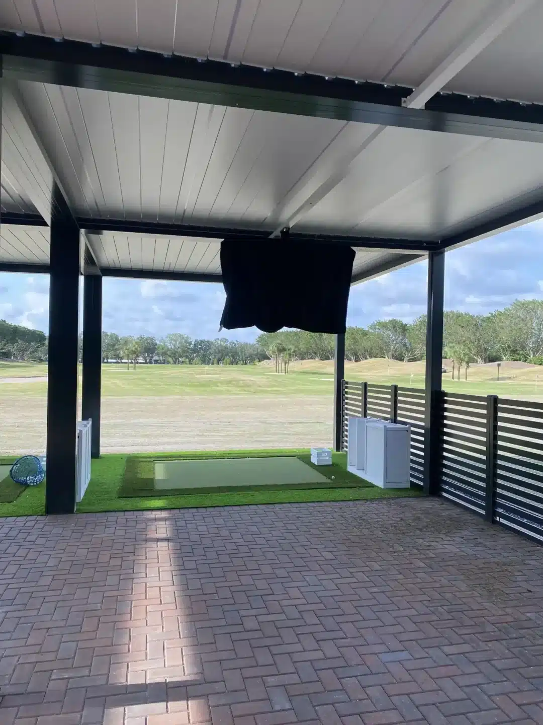 Covered outdoor patio with white metal roof and black fabric hanging from beam; view of a grassy golf course beyond, brick-paved foreground and white storage boxes nearby.