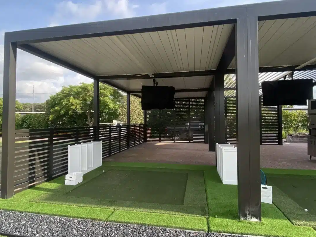 OPEN-air pavilion with dark grey metal framework and white slatted ceiling over a paved area with green artificial turf and black horizontal fencing.