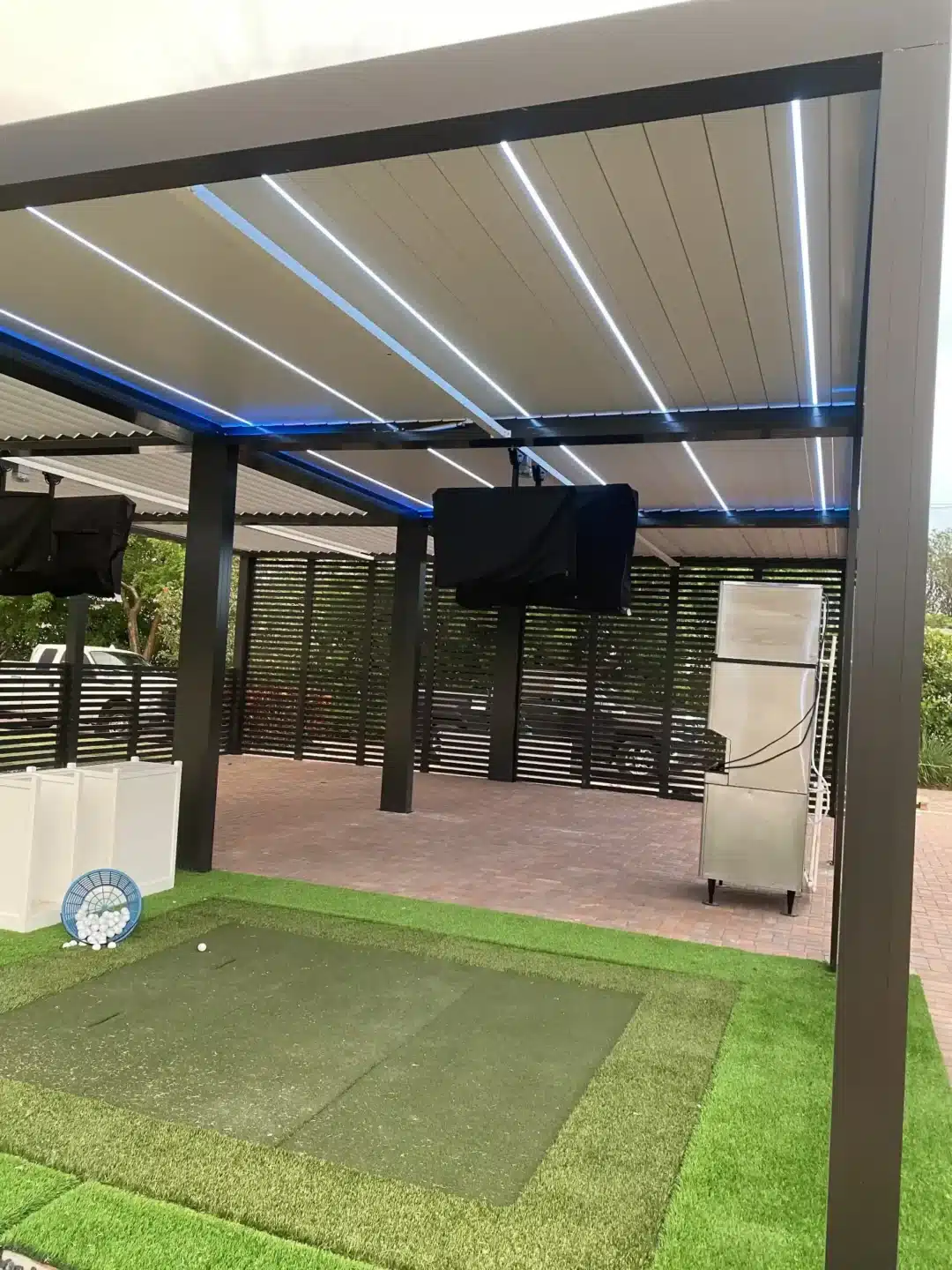 Outdoor covered patio with overhead LED light strips and mounted screens for entertainment, on a black metal frame structure.