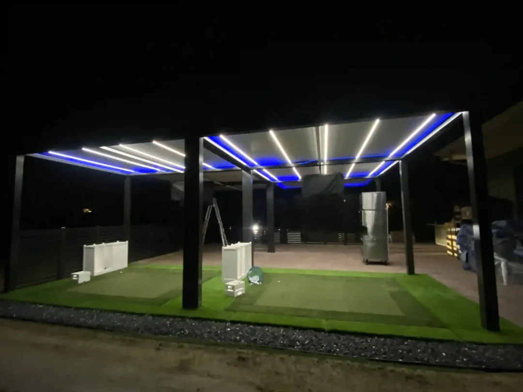 Night view of an outdoor covered patio at night with blue and white LED strip lighting, scattered boxes and equipment on artificial turf.