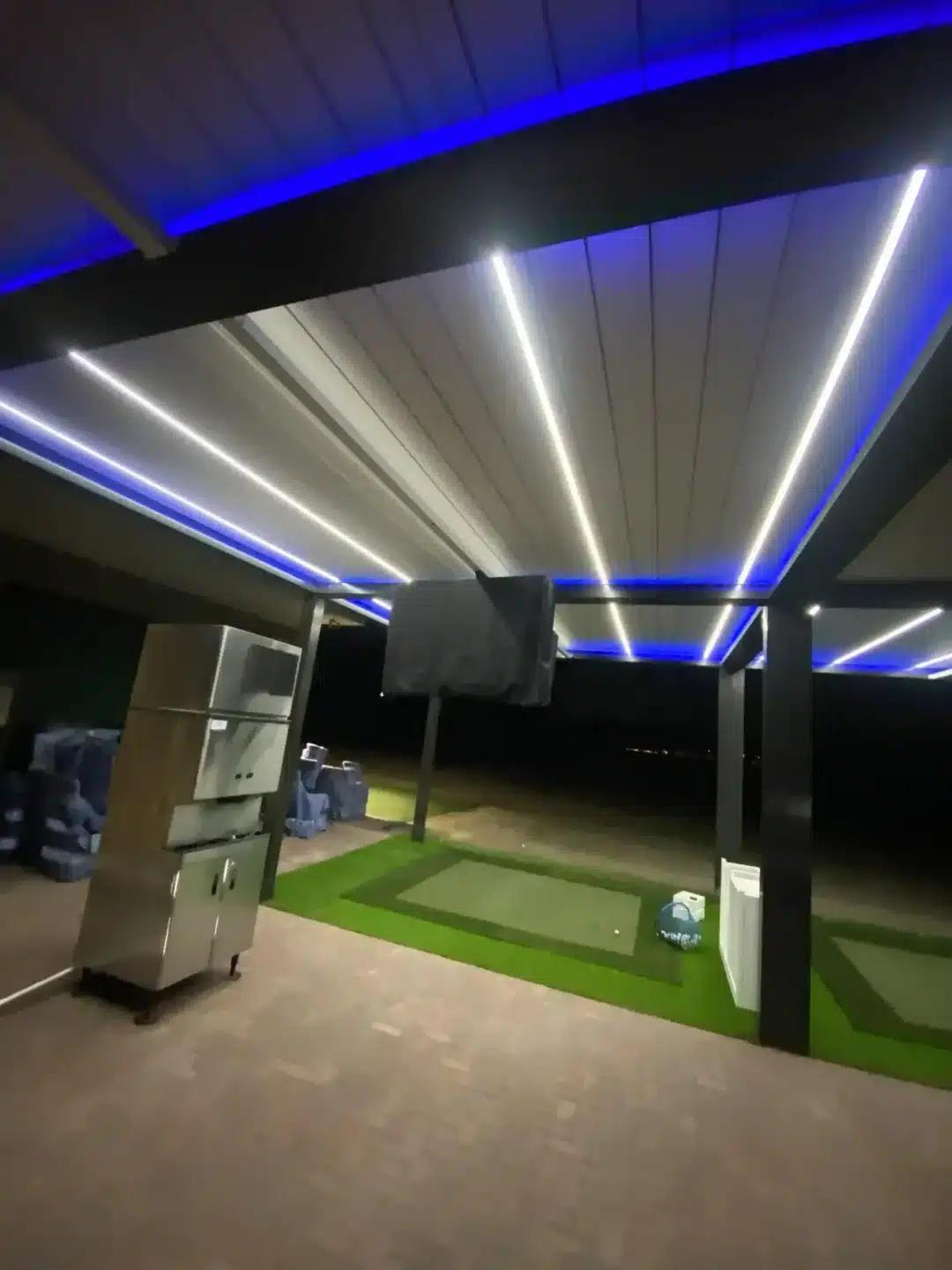 Indoor space with blue neon ceiling lights, dark pillars, and a small artificial turf putting area