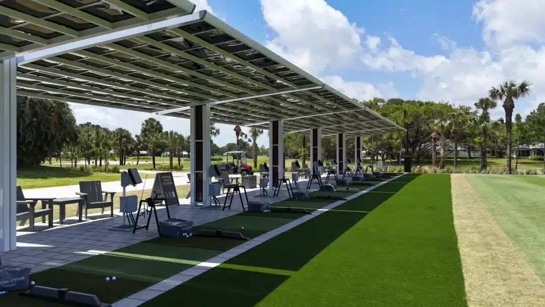 Golf driving range with covered bays, hitting mats, and ball baskets under a metal roof, palm trees in background under a blue sky