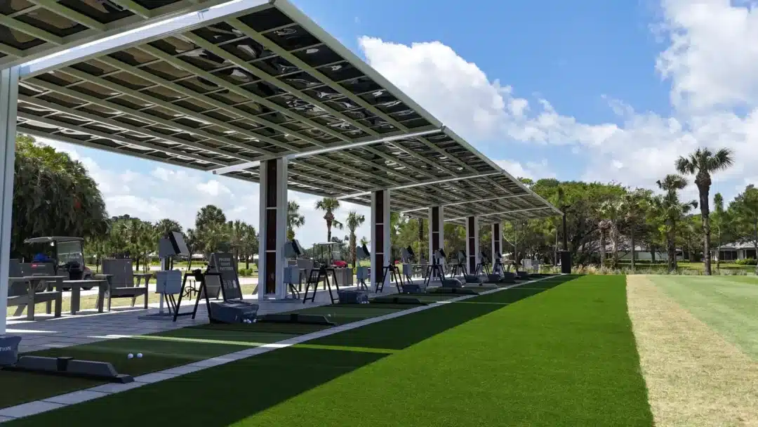 Row of golf practice bays under a metal canopy at a sunny driving range with palm trees in the background.