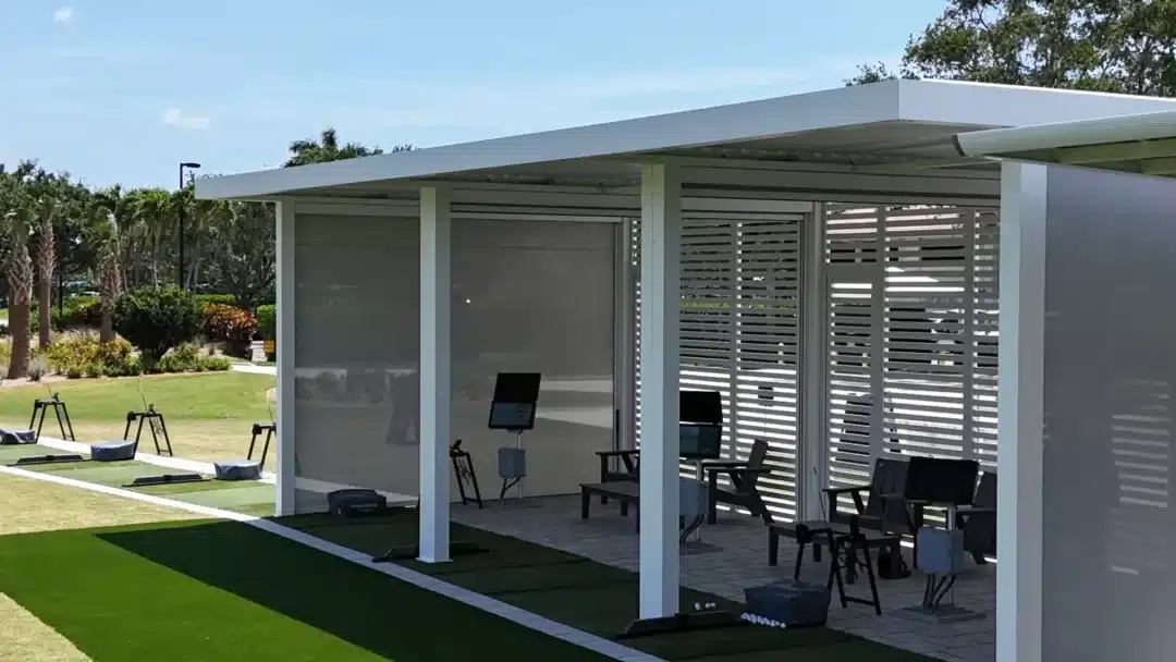 Shaded outdoor golf practice area with seating, screens, and hitting mats under a modern white shelter.