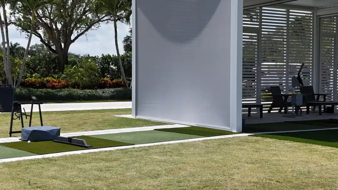 Outdoor lawn beside a modern pavilion with horizontal slats and seating inside; a small golf practice area with mats and a ball in the foreground
