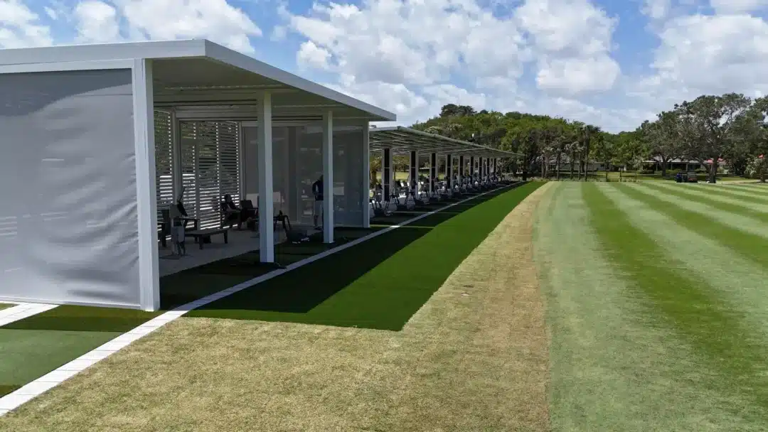 Row of shade-covered bays along a golf driving range with people working at tables under a modern white shelter.