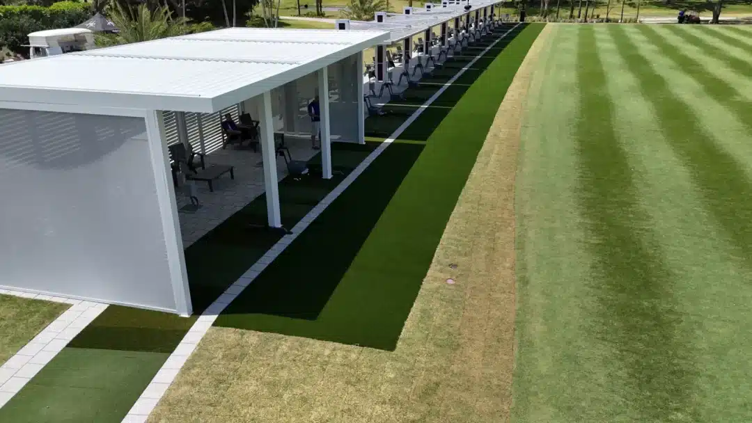 Aerial view of a golf practice range with a long row of covered bays, mats, and a striped green outfield.” (informative)