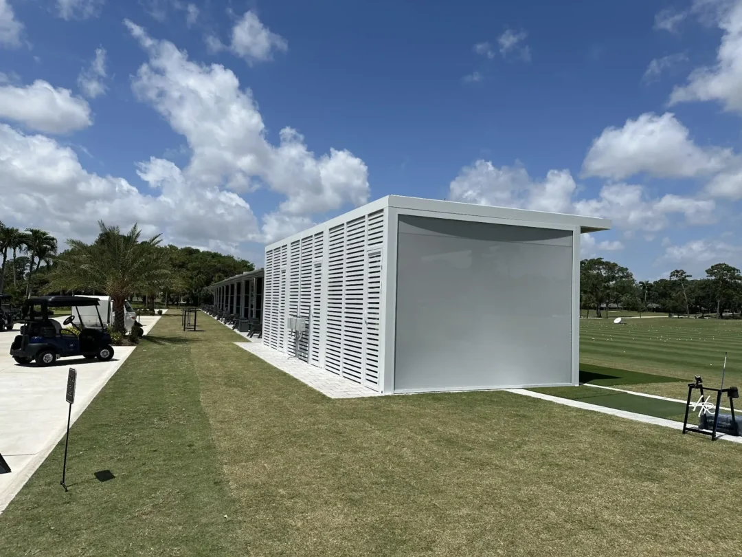 Modern white utility building with louvered walls beside a row of golf carts under a blue sky with clouds.