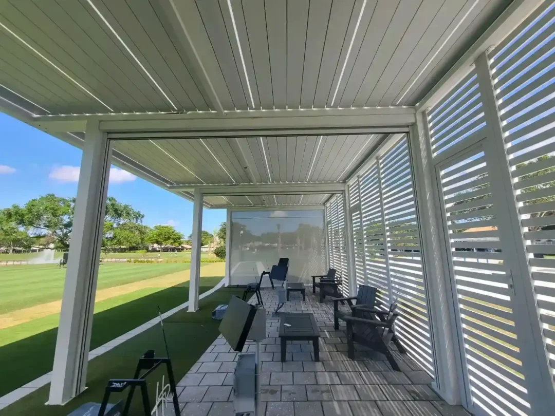Covered outdoor seating area under a white metal canopy with slatted walls, multiple chairs and small tables on a paved patio overlooking a green lawn and clear blue sky.
