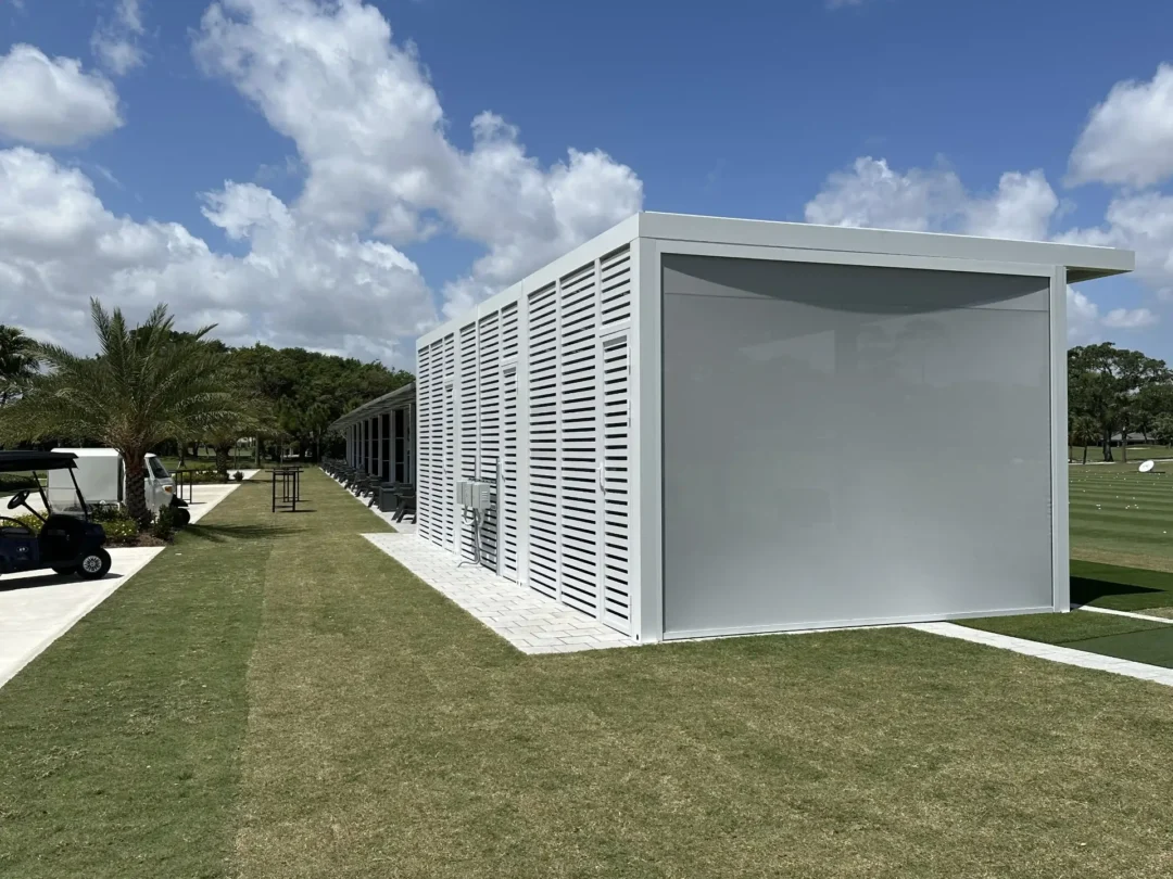 Modern white slatted building along a paved path, with palm trees and a blue sky above scattered with clouds.