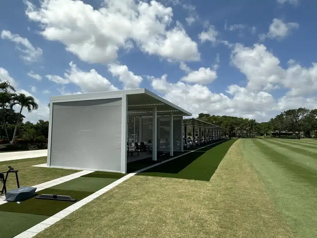 Row of covered golf practice bays with artificial turf, under a bright blue sky with scattered clouds