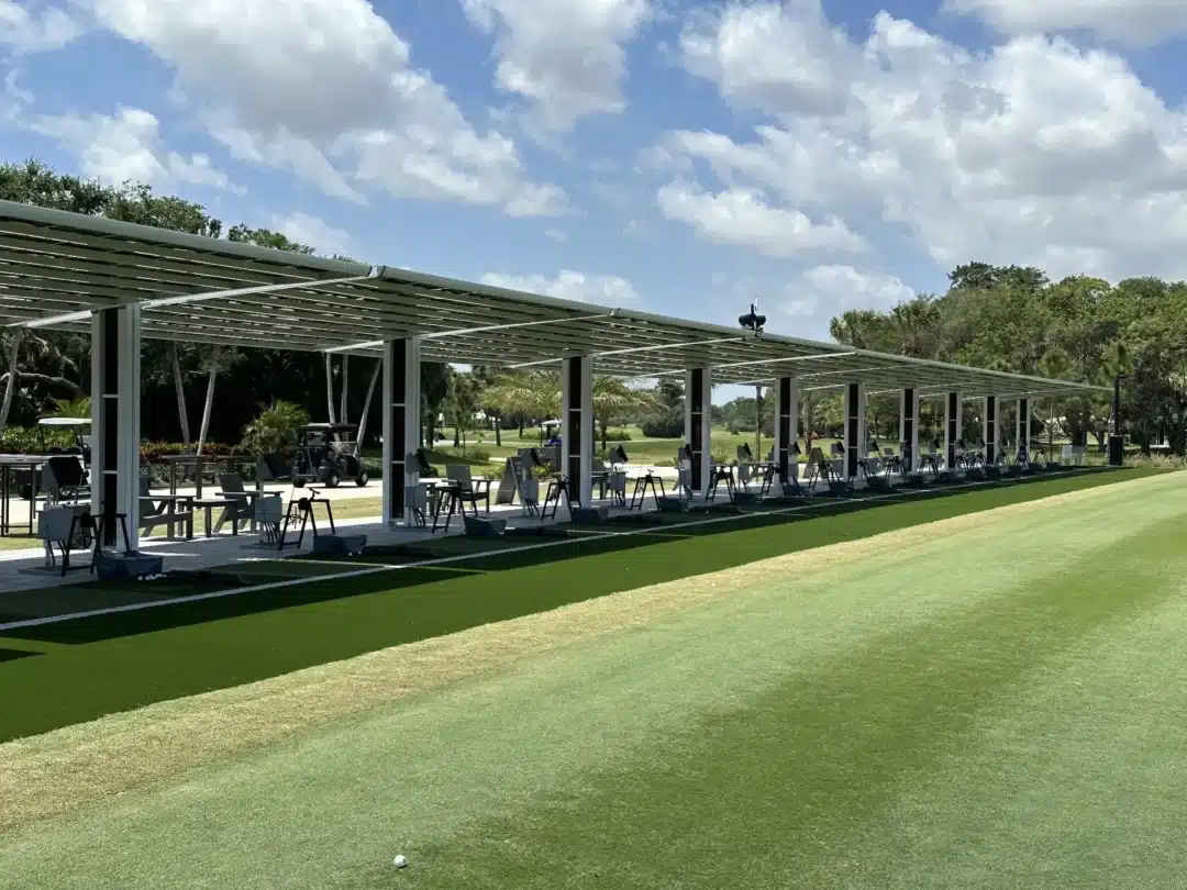 Golf driving range with multiple open bays under a metal canopy, rows of chairs and golf equipment, a lone ball on the grass in the foreground, under a blue sky.