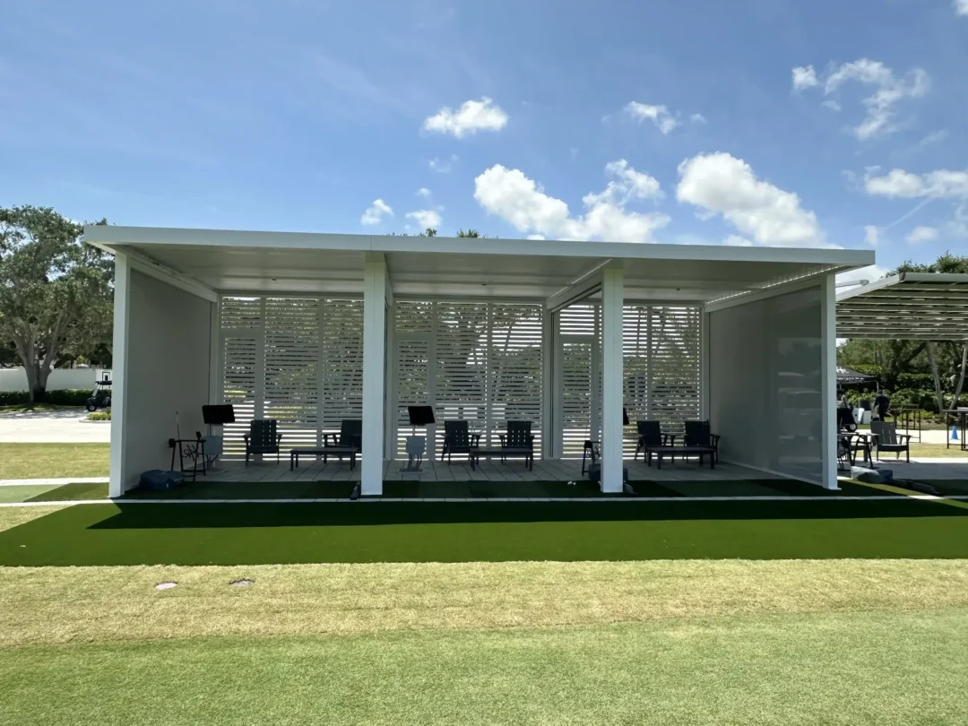Open-air white pavilion with slatted walls and multiple chairs arranged inside, under a clear blue sky on a sunny day.