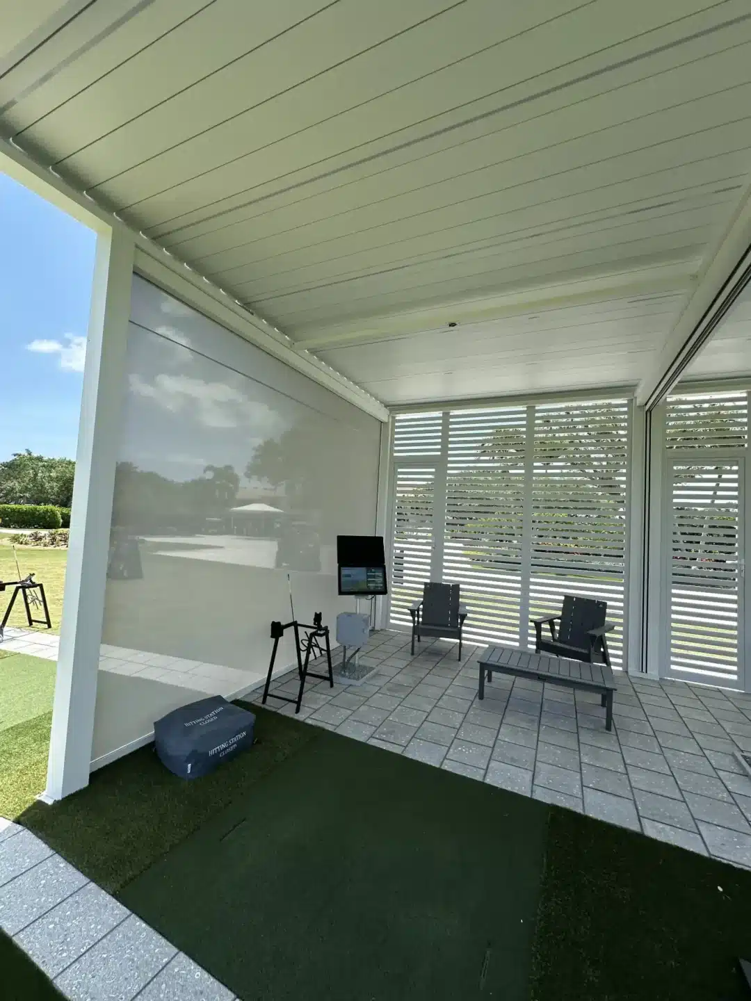 Covered outdoor patio with white slatted walls, grey seating, and a coffee table on tiled floor; artificial turf at the edge.