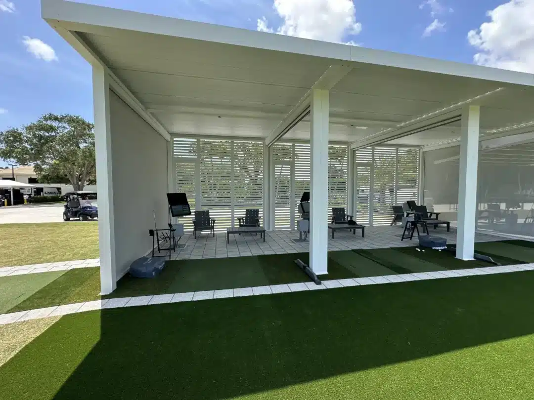 Outdoor white pavilion for golf practice with seating, slatted walls, and artificial turf mats.