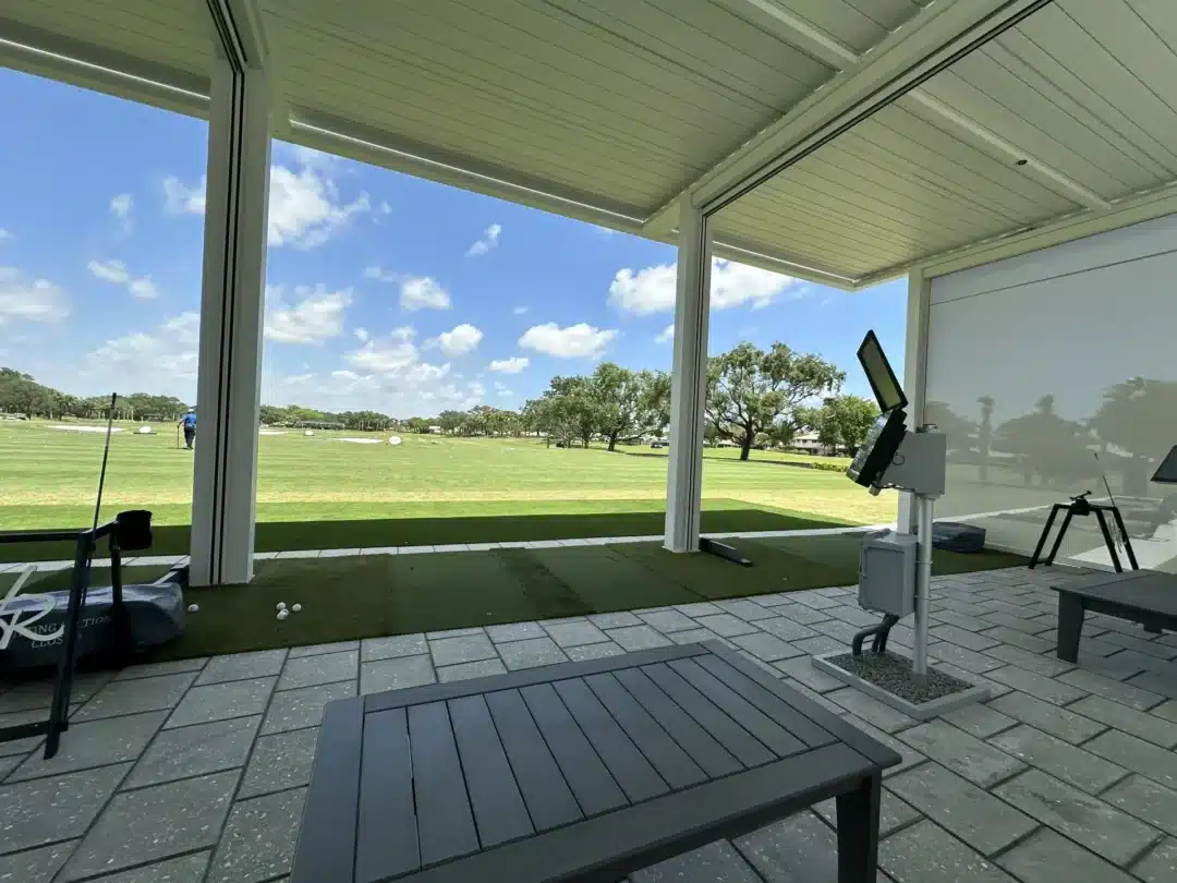 Covered golf practice bay with turf mats, a monitor on a stand, and scattered golf balls near a bench; open field and trees beyond.