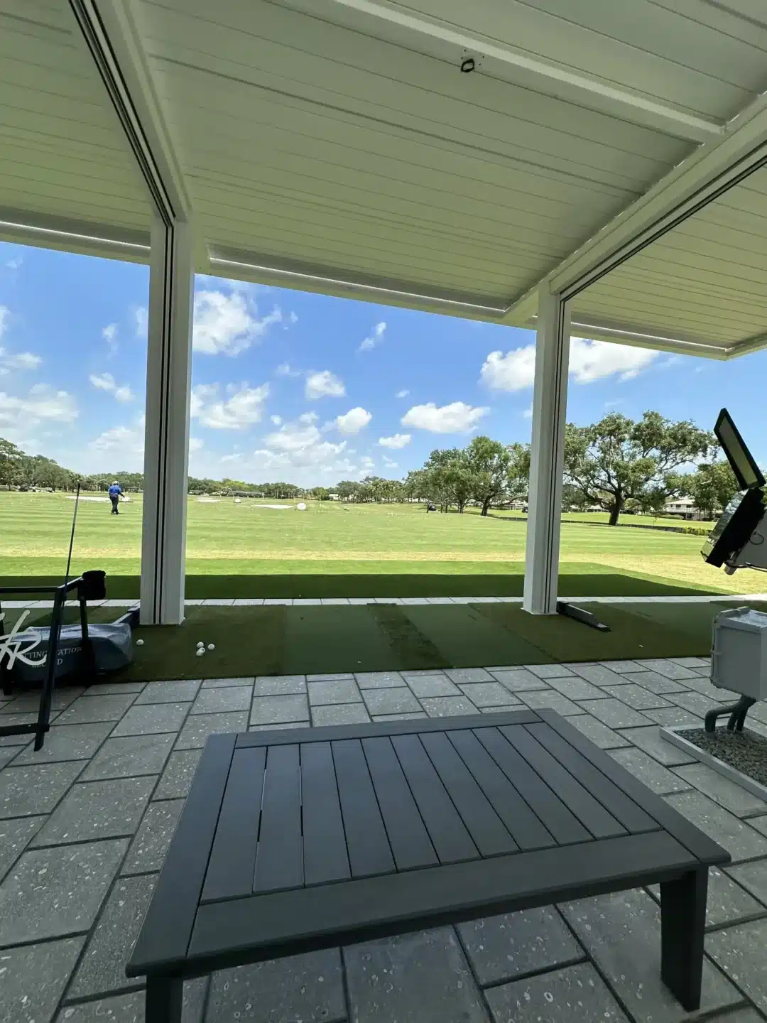 Covered outdoor seating area overlooking a grass driving range with a person practicing in the distance; a table in the foreground and a monitor on the right.