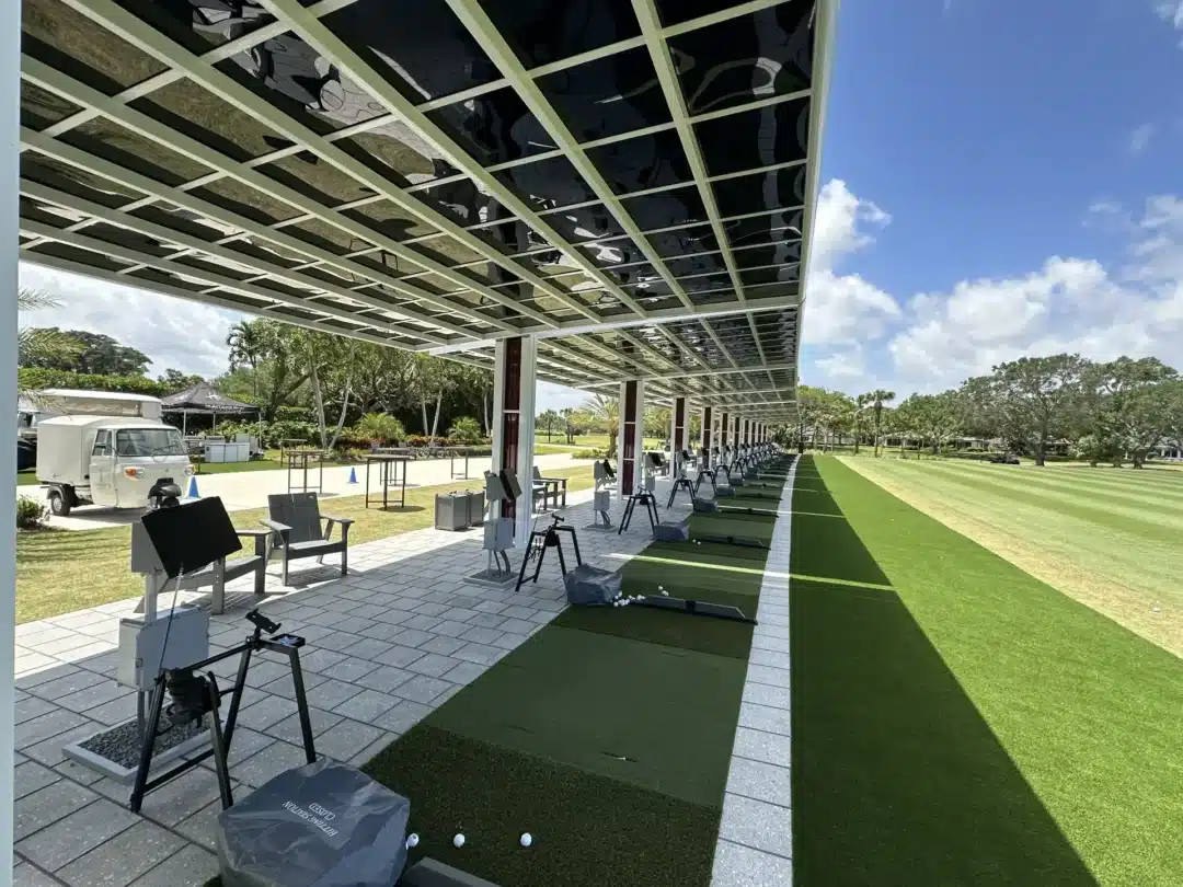 Outdoor golf practice area with a long covered bays and mats, with chairs and golf clubs set up; blue sky and trees in the background