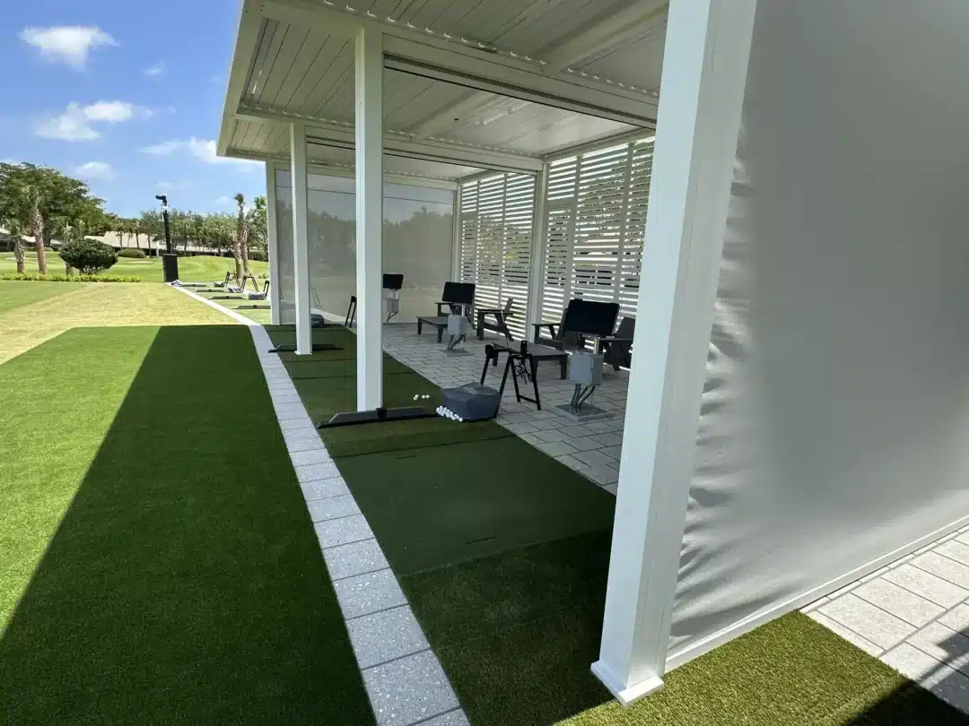Outdoor golf practice area with artificial turf mats under a white covered walkway and a row of chairs inside a glass-walled shelter.