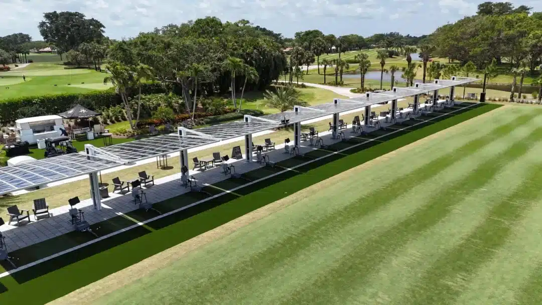A golf driving range with a long row of covered practice bays, chairs and tables, and palm trees in the background under a sunny sky.