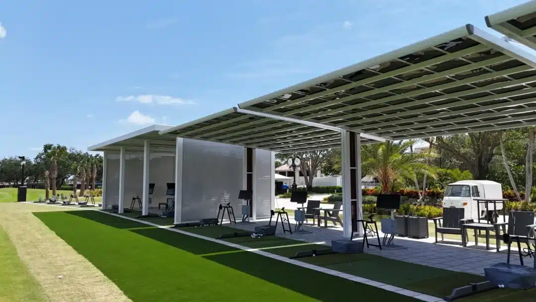 Outdoor golf driving range with a covered practice area, artificial turf mats, and seating under a modern canopy.