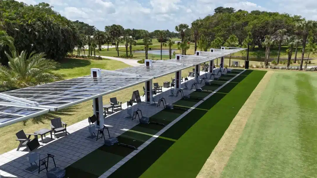 Driving range with row of covered bays, turf mats, and seating under a glass-roof shelter, palm trees in the background.