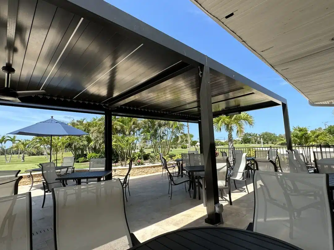 Outdoor patio with metal pergola, glassy patio chairs, and tables under a sunny blue sky; palm trees in the background.
