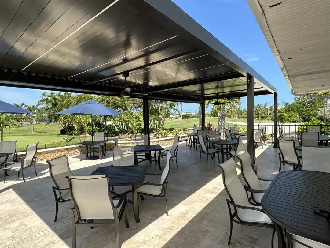 Outdoor restaurant patio under a dark metal canopy with black tables and cream chairs, white umbrellas, and a sunny green lawn beyond.