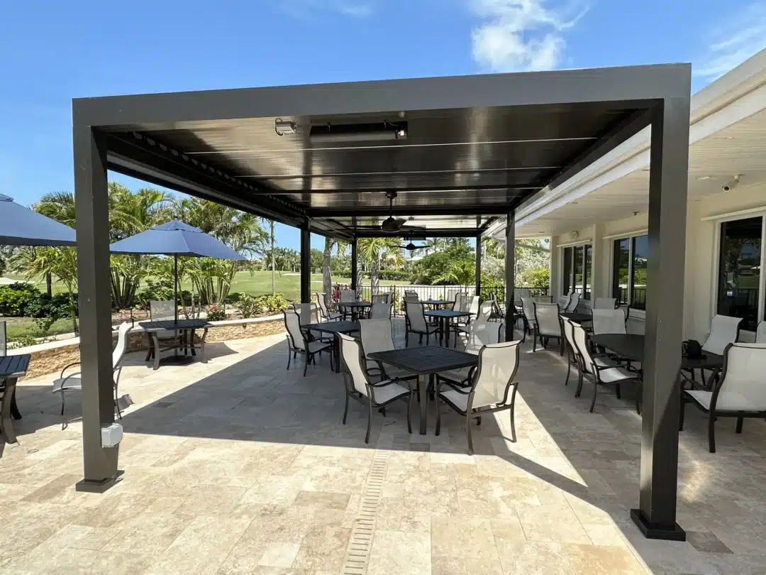 Outdoor patio with black metal pergola, several tables and cream chairs under a covered seating area; blue sky and palm trees in background.