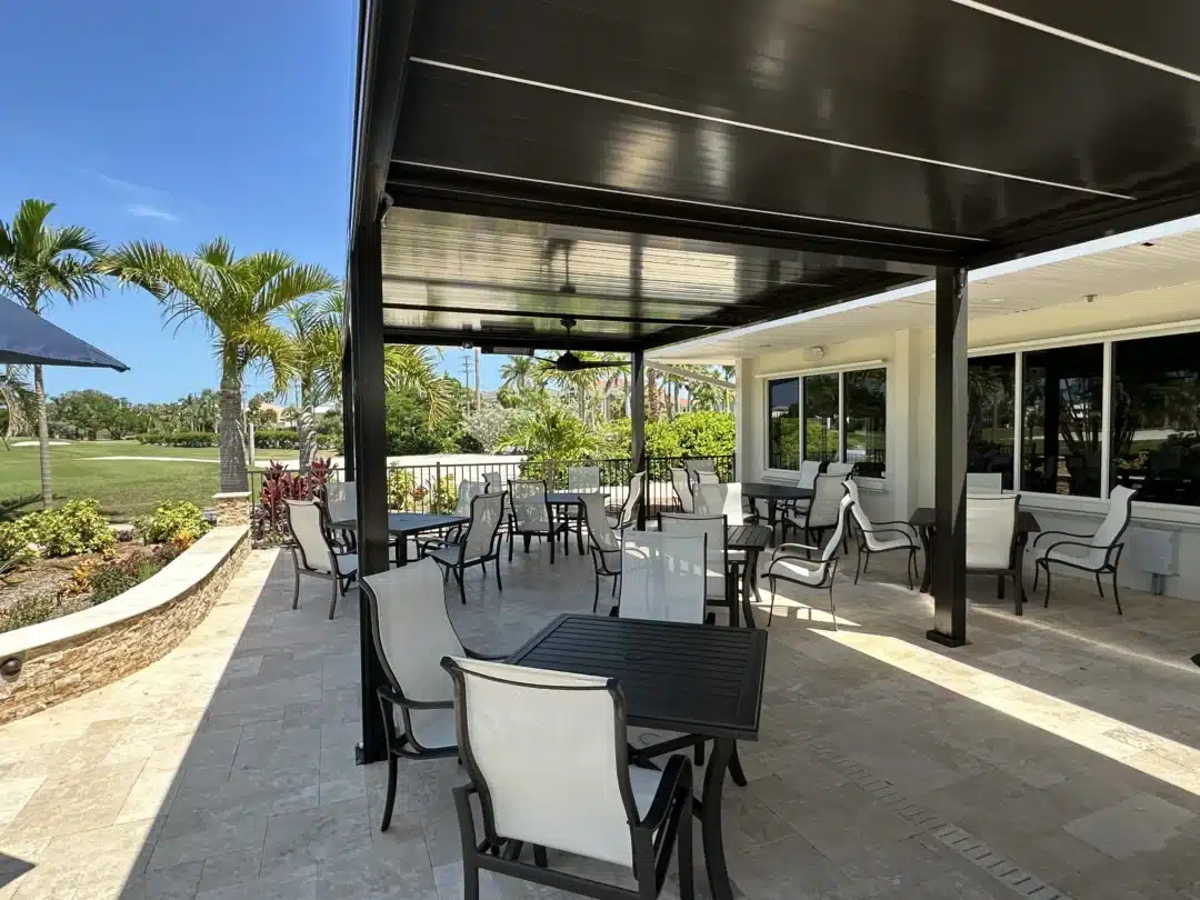 Outdoor patio seating area with black metal tables and white chairs under a dark canopy, palm trees and blue sky in the background.