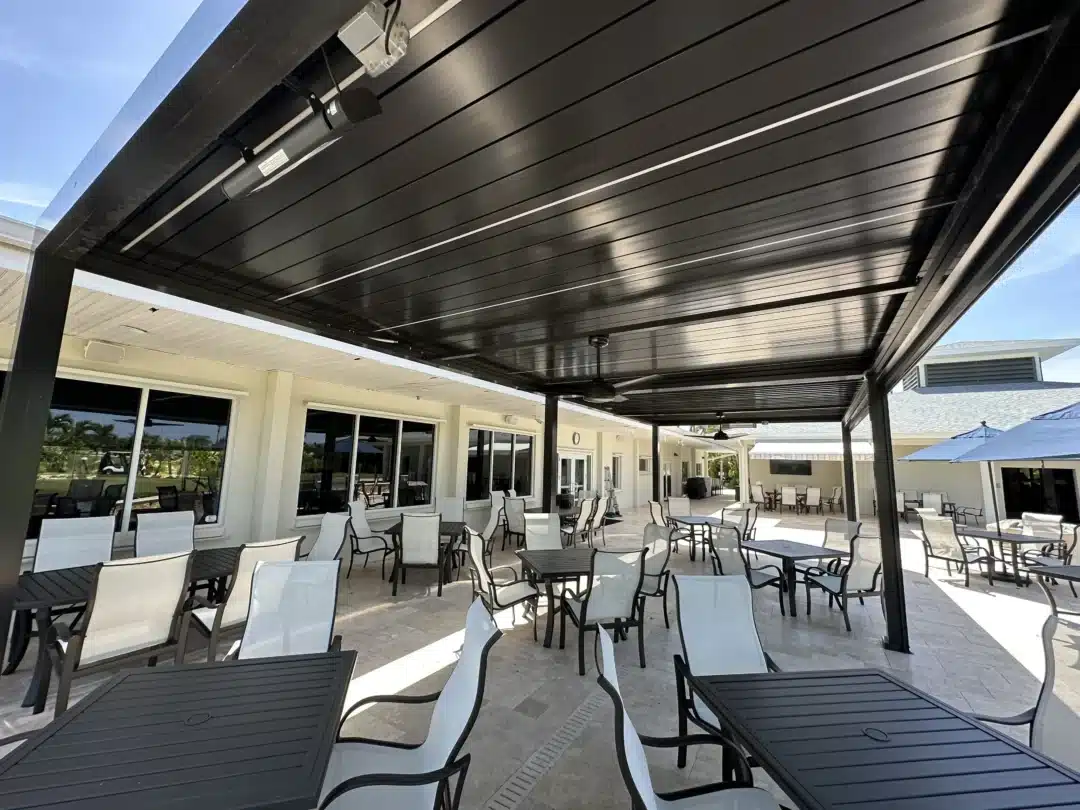 Outdoor patio under a dark metal slatted roof with many black tables and white chairs. The glass-walled building and blue sky are in the background.