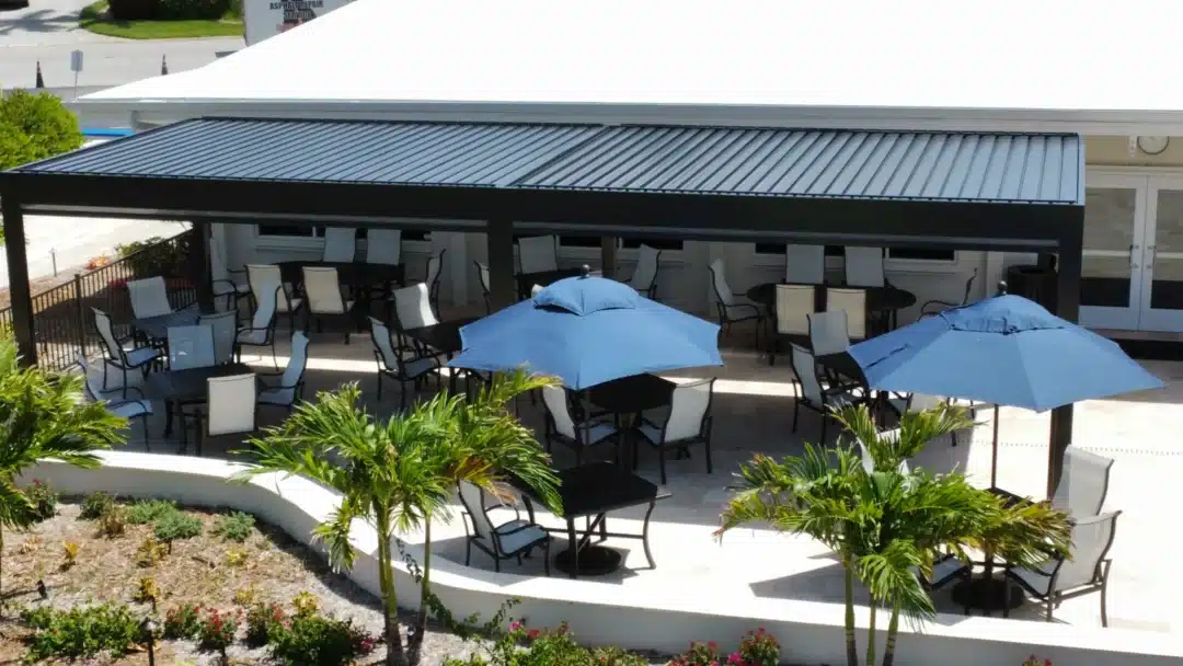 Outdoor patio with black metal tables and white chairs under a dark canopy; blue umbrellas, palms, and surrounding landscaping.