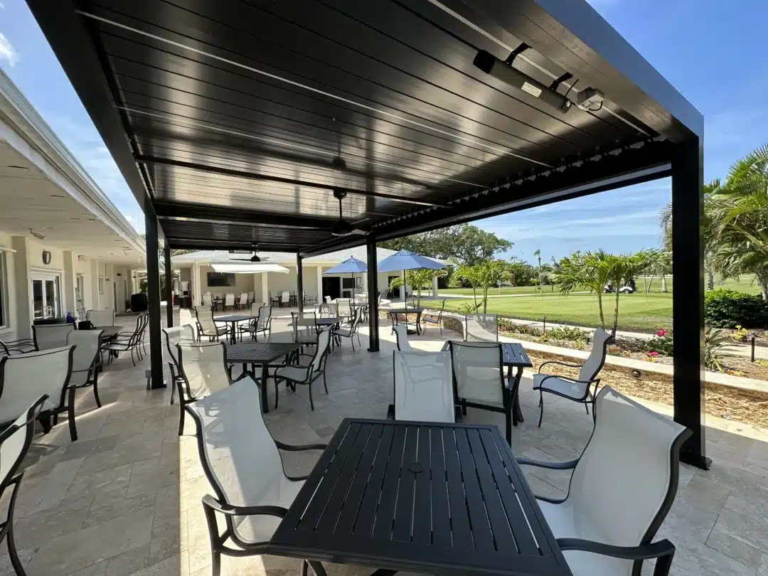 Outdoor covered patio with black metal tables and white chairs under a dark metal canopy, sunny day with palm trees in the background.