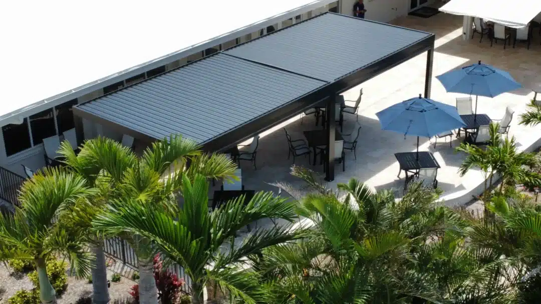 Outdoor patio area with blue umbrellas, tables, and chairs under a metal-roofed canopy, framed by palm trees and tropical landscaping.
