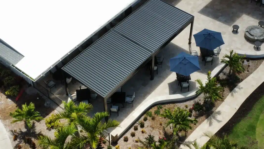 Aerial view of a patio with blue umbrellas, tables and chairs under a metal slatted roof, surrounded by palm trees and stone pathways.