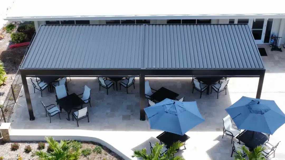 Bird's-eye view of an outdoor dining area under a metal-roofed pavilion with multiple tables, chairs, and blue umbrellas on a tiled patio.