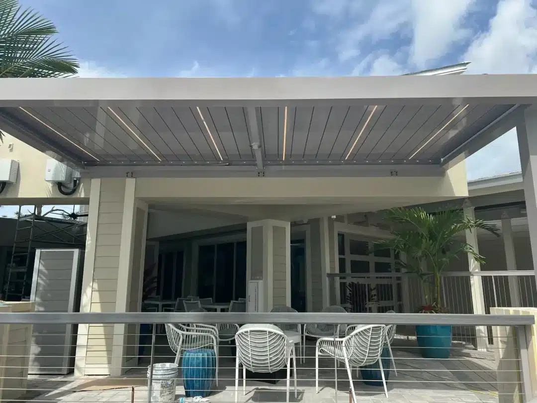 Outdoor patio under a modern slatted roof with white chairs around a table, railing, and potted palms near a light building under a blue sky.