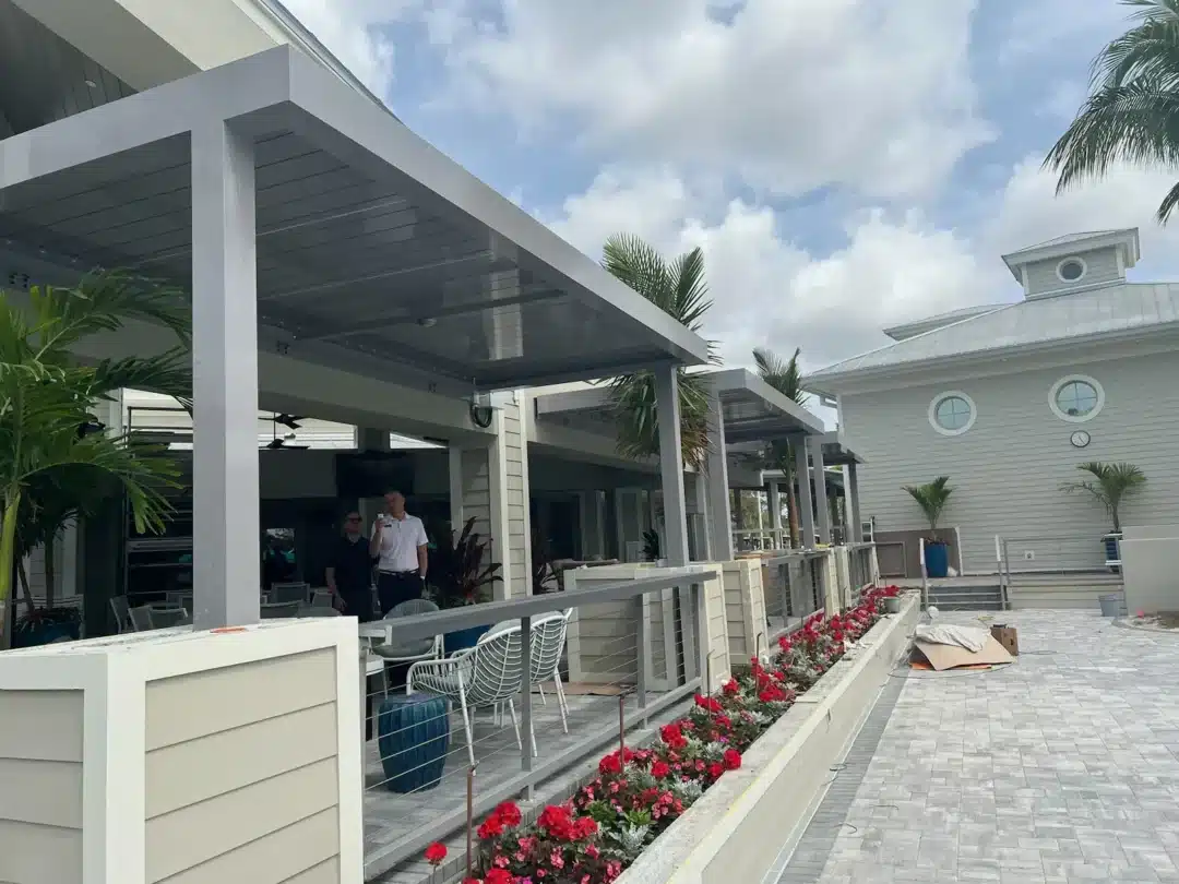 Two men stand under a gray pergola on a shaded patio with white railings and outdoor chairs, next to a row of red flowers in planters.