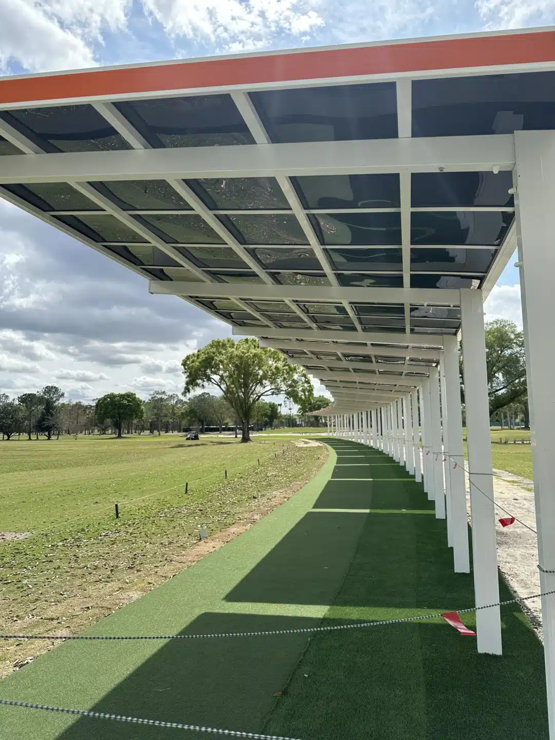Covered walkway with white posts and glass-panel canopy extending along a green artificial turf path under a blue, partly cloudy sky.