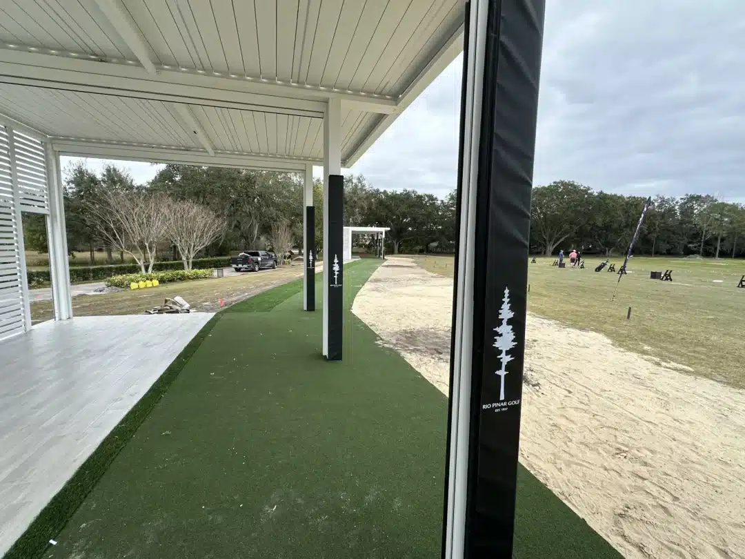 Covered golf driving range with white overhead canopy and green artificial turf; black pillars with tree logos mark hitting bays, distant players practicing.